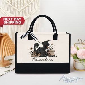 May include: A black and white tote bag featuring a black dragon design and the name Alexandrina printed on the front. The bag has black handles and a black base.