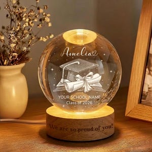 May include: A clear crystal ball night light with a graduation cap and diploma design. The text "Amelia", "YOUR SCHOOL NAME", and "Class of 2026" are engraved on the crystal. The wooden base reads "We are so proud of you!"
