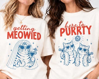 Op de afbeelding: Twee off-white T-shirts met blauwe en rode grafische prints. Het linker shirt toont "getting MEOWIED" met een kat in een sluier met een martini. Het rechter shirt toont "here to PURRTY" met twee katten met zonnebrillen onder een discobal.