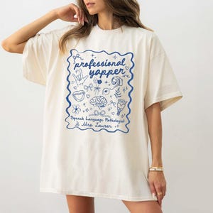 May include: A white oversized t-shirt with a blue graphic print that says "professional yapper" and includes various illustrations. The text below the graphic says "Speech Language Pathologist Mrs. Lauren."