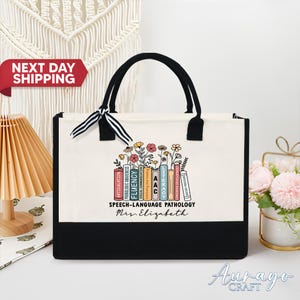 May include: A canvas tote bag with black handles and a black base. The bag displays a colourful graphic of books and flowers with the text "SPEECH-LANGUAGE PATHOLOGY Mrs. Elizabeth."