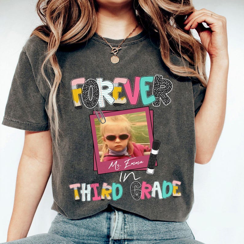 May include: A charcoal gray t-shirt featuring the text FOREVER in colorful block letters, a pink framed photograph of a child wearing sunglasses, and the text Ms. Emma in THIRD GRADE below the photo, accented with a paperclip and pencil graphic.