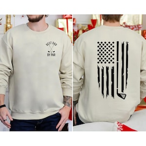 Peut inclure: Un sweat-shirt beige avec un motif sur le th&egrave;me du golf. Le devant du sweat-shirt porte le texte "Best Dad By Par" avec un graphique de club de golf et de balle. Le dos du sweat-shirt pr&eacute;sente un motif de drapeau am&eacute;ricain d&eacute;labr&eacute; avec un club de golf et des balles de golf.