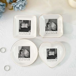May include: Four white ceramic trinket dishes with gold-toned rims, each featuring a black and white photograph and the printed text "Be the things you loved the most about her" with a small heart symbol at the end of the phrase.