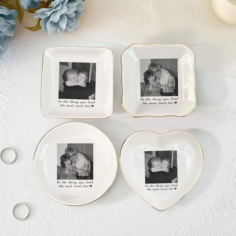 May include: Four white ceramic trinket dishes with gold-toned rims, each featuring a black and white photograph and the printed text "Be the things you loved the most about her" with a small heart symbol at the end of the phrase.