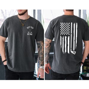May include: A dark gray t-shirt with a white graphic on the front that says "Best Dad By Par" with a golf club design. The back of the shirt features a white distressed American flag design with a golf club.