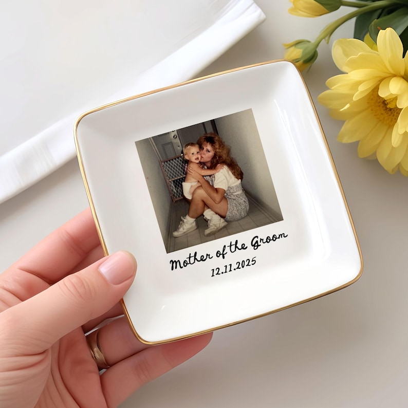 Op de afbeelding: Een witte keramische sieradenschaal met een gouden rand. De schaal heeft een foto van een moeder en kind. De tekst "Mother of the Groom 12.11.2025" staat op de schaal. De schaal wordt door een hand vastgehouden.