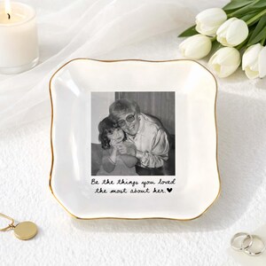 May include: A square white ceramic trinket dish with gold-trimmed edges. The centre features a black and white photograph of an adult and a child, with the text "Be the things you loved the most about her." printed in black script below the image.