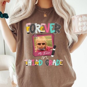 May include: A brown t-shirt featuring a graphic with the text FOREVER IN THIRD GRADE. The design includes a pink photo frame containing a picture of a child wearing sunglasses, a paperclip, a pencil, and the handwritten name Ms. Emma.