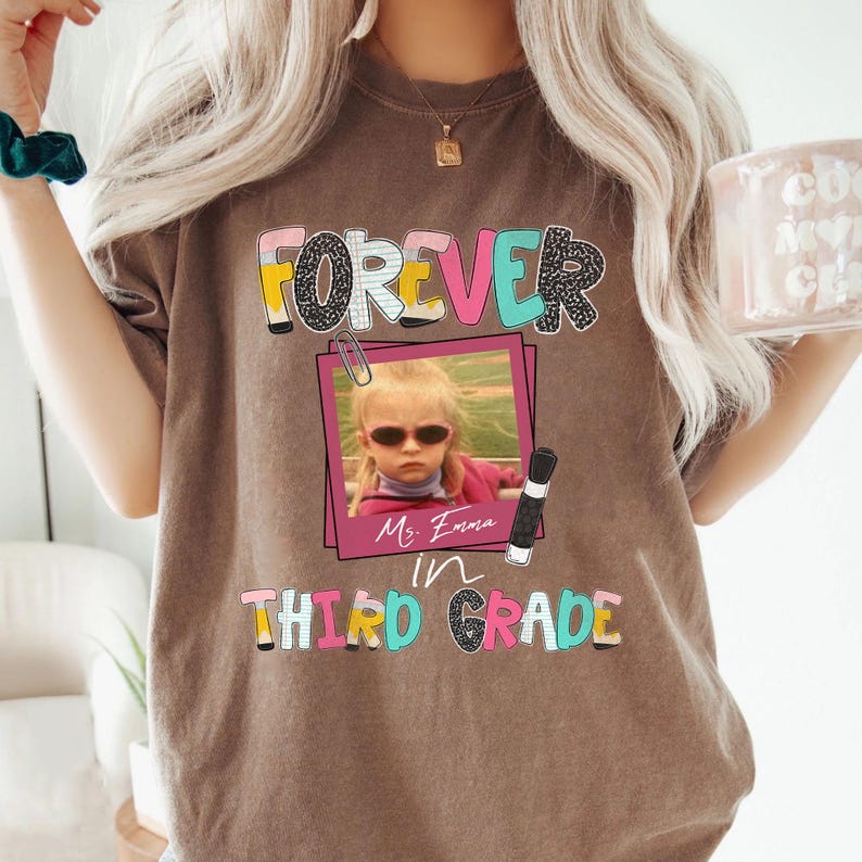 May include: A brown t-shirt featuring a graphic with the text FOREVER IN THIRD GRADE. The design includes a pink photo frame containing a picture of a child wearing sunglasses, a paperclip, a pencil, and the handwritten name Ms. Emma.
