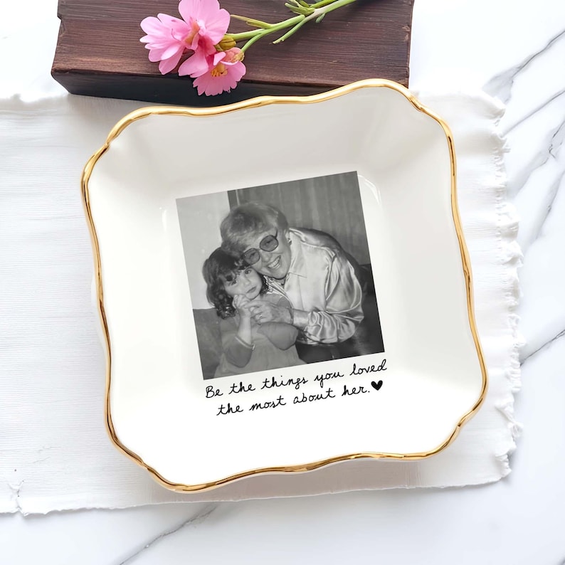 May include: A square white ceramic trinket dish with a scalloped gold-painted rim. The centre features a black and white photograph of an adult and a child, with the text "Be the things you loved the most about her." printed in black script below.