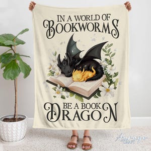 May include: A cream-coloured fleece blanket with the text "In a world of bookworms be a book dragon" in black lettering. The blanket features an illustration of a black and yellow dragon curled up on an open book.