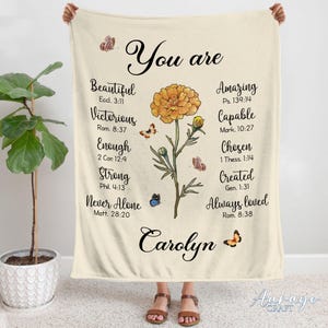 May include: A beige fleece blanket featuring a floral design and the text "You are" with a list of positive affirmations and bible verses. The name "Carolyn" is written in cursive at the bottom of the blanket.