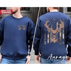 May include: Navy blue sweatshirt with a deer graphic on the back, featuring a camouflage and American flag design. The front has a small gold emblem with the letters "DD". The text "NEXT DAY SHIPPING" is visible.