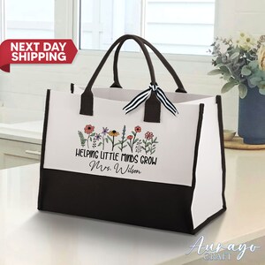 May include: A white and black tote bag with a black handle and a black and white ribbon. The bag has a white panel with a floral design and the text "Helping Little Minds Grow Mrs. Wilson".