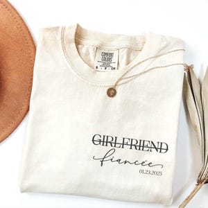 May include: A beige t-shirt with the text "GIRLFRIEND fianc&eacute;e 01.23.2025" printed on the front.