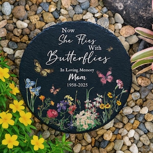 May include: A circular, black memorial stone inscribed with "Now She Flies With Butterflies" and "In Loving Memory Mom 1958-2025". It features colourful butterflies and flowers. The stone is set amongst pebbles and greenery.