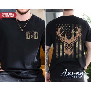 May include: Black t-shirt featuring a deer design. The front of the shirt displays the word "DAD" in a camouflage pattern. The back of the shirt showcases a deer head with antlers set against an American flag design with a camouflage pattern.