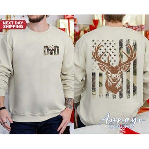 May include: A beige sweatshirt with a deer-themed design. The front features the word "DAD" with a deer silhouette. The back displays a deer head with antlers, set against a camouflage and American flag design. The sweatshirt has long sleeves.