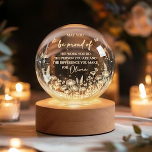 May include: A clear glass sphere resting on a cylindrical wooden base. The sphere features etched floral designs, butterflies, and the text: MAY YOU be proud of THE WORK YOU DO THE PERSON YOU ARE AND THE DIFFERENCE YOU MAKE FOR Olivia.