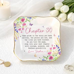May include: A white ceramic trinket dish with a gold rim, adorned with floral and butterfly designs. The dish features the text "Chapter 30" and a personalized message. A candle, flowers, and jewelry are in the background.