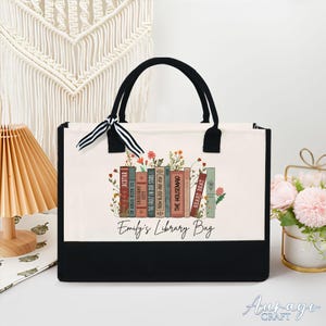May include: A white canvas tote bag with black trim and handles. The bag features a design of stacked books with floral accents and the text "Emily's Library Bag".