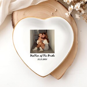 Op de afbeelding: Wit hartvormig keramisch schaaltje met een gouden rand. Het schaaltje heeft een foto van een moeder en kind, met de tekst "Mother of the Bride" en de datum "12.11.2025". Het schaaltje staat op een houten schijf.