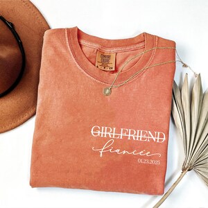 May include: A coral-colored t-shirt with the text "GIRLFRIEND fiancee 01.23.2025" printed in white.
