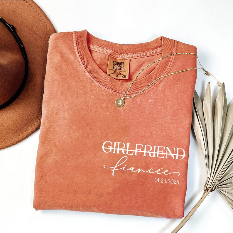 May include: A coral-colored t-shirt with the text "GIRLFRIEND fiancee 01.23.2025" printed in white.