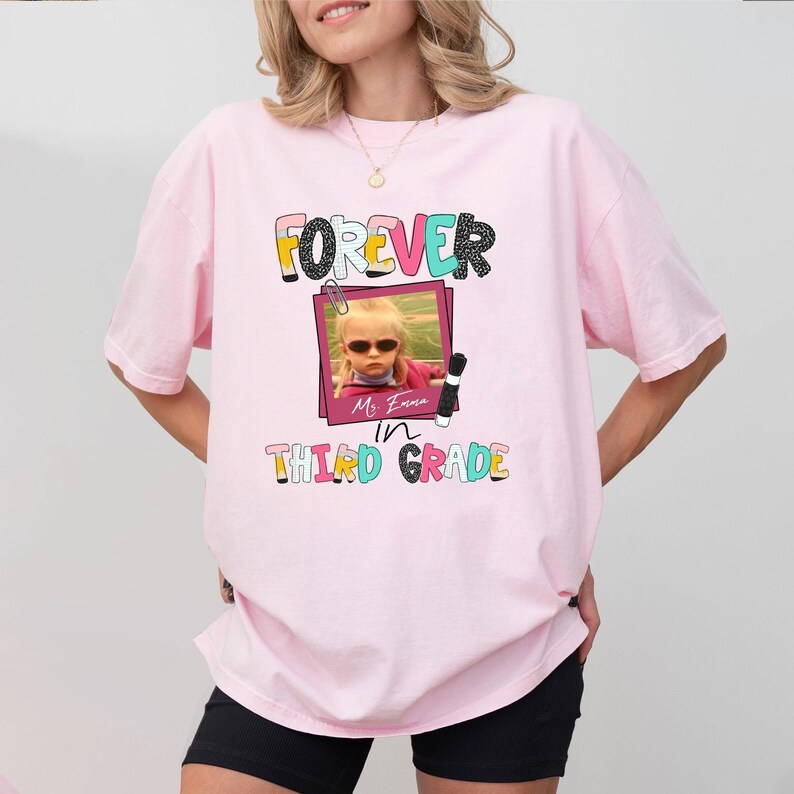 May include: A light pink t-shirt featuring a graphic print with the text FOREVER IN THIRD GRADE. The design includes a framed photograph of a child wearing sunglasses, a paperclip icon, a pencil, and colorful, patterned lettering.