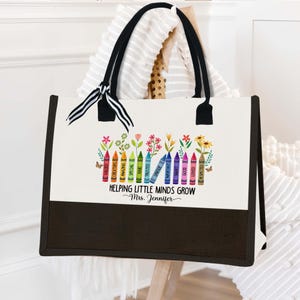 Puede incluir: Una bolsa de lona blanca con asas negras y fondo negro. La bolsa tiene un dise&ntilde;o colorido de l&aacute;pices de colores con palabras como "valued", "creators", "amazing", "special", "kind", "trusted", "unique", "important", "smart", "brave", "good", "loved", "respected" impresas en ellos. El texto "Helping Little Minds Grow" y "Mrs. Jennifer" est&aacute; impreso debajo de los l&aacute;pices de colores.