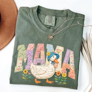 May include: Olive green Comfort Colors t-shirt with the word "MAMA" in patchwork letters. A cartoon duck wearing a blue hat is featured below the word. A gold necklace is draped across the shirt.