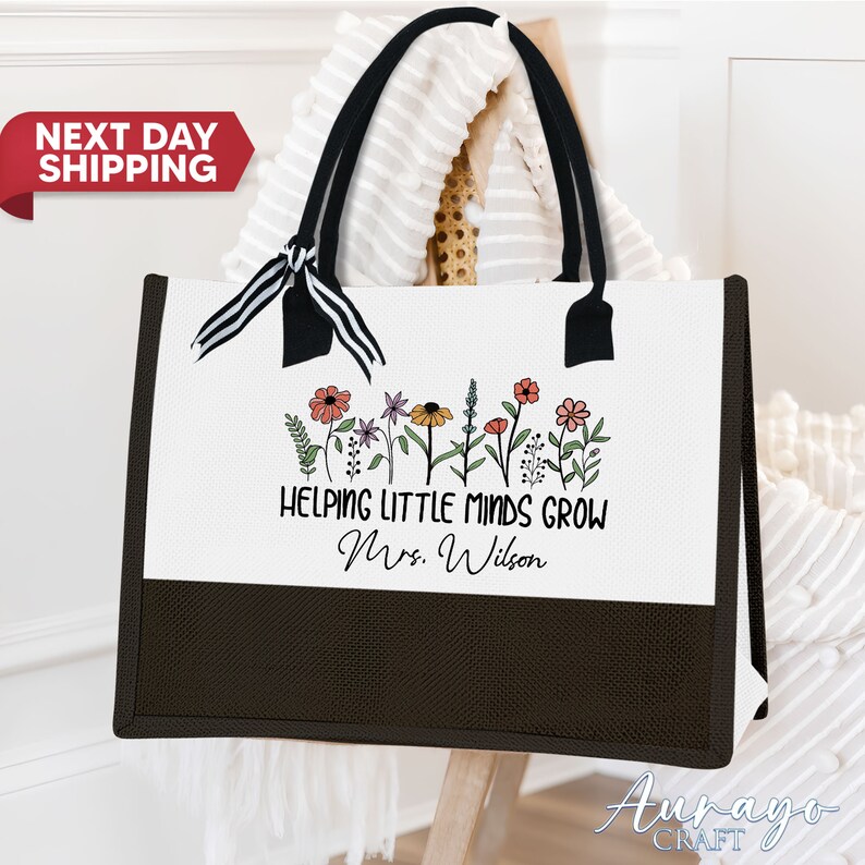 May include: White canvas tote bag with black handles and a black and white ribbon. The bag has a black bottom and features a line of colorful flowers and the text "Helping Little Minds Grow Mrs. Wilson".