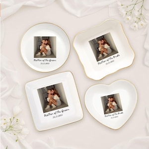 Op de afbeelding: Vier witte keramische sieradenbakjes met gouden randen, elk met een foto en tekst. De vormen zijn een cirkel, een vierkant, een hart en een unieke vorm. Tekst luidt "Mother of the Groom" of "Mother of the Bride" met een datum.