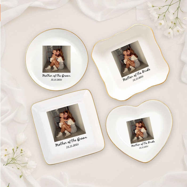 Op de afbeelding: Vier witte keramische sieradenbakjes met gouden randen, elk met een foto en tekst. De vormen zijn een cirkel, een vierkant, een hart en een unieke vorm. Tekst luidt "Mother of the Groom" of "Mother of the Bride" met een datum.