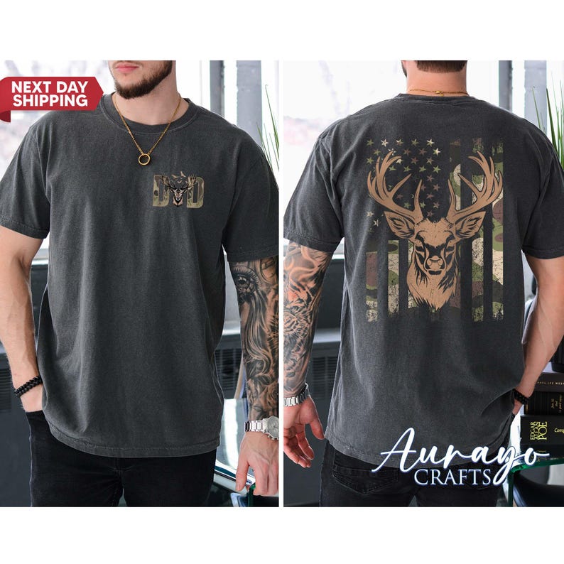 May include: Dark gray t-shirt featuring a deer head design with an American flag motif on the back. The front of the shirt has the word "DAD" in a camouflage design. The shirt is short-sleeved.
