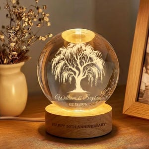 May include: A clear glass sphere with a willow tree design, illuminated from within, sits on a wooden base. The base reads "HAPPY 50TH ANNIVERSARY". The sphere is engraved with the names "William & Sophia" and the date "02.15.1976".