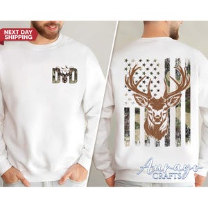 May include: White sweatshirt with a deer and camouflage design. The front features the letters "DAD" with a deer head graphic. The back displays a deer head over a camouflage American flag design. The sweatshirt has a crew neck.