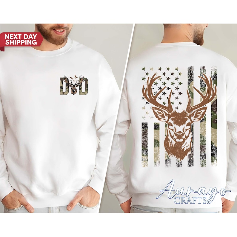 May include: White sweatshirt with a deer and camouflage design. The front features the letters "DAD" with a deer head graphic. The back displays a deer head over a camouflage American flag design. The sweatshirt has a crew neck.