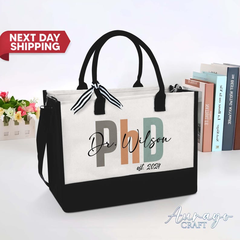 Pode incluir: Uma bolsa tote preta e branca com um painel de lona branca que apresenta o texto "Dr. Wilson PhD est. 2024" em letras coloridas. A bolsa tem uma al&ccedil;a preta e al&ccedil;as pretas.