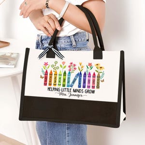 Puede incluir: Una bolsa de lona blanca con asas negras y fondo negro. La bolsa tiene un dise&ntilde;o colorido de l&aacute;pices de colores con palabras como "valued", "creators", "amazing", "special", "kind", "trusted", "unique", "invincible", "smart", "important", "good", "loved" y "respected". El texto "Helping Little Minds Grow" est&aacute; impreso debajo de los l&aacute;pices de colores, seguido de "~Mrs. Jennifer".