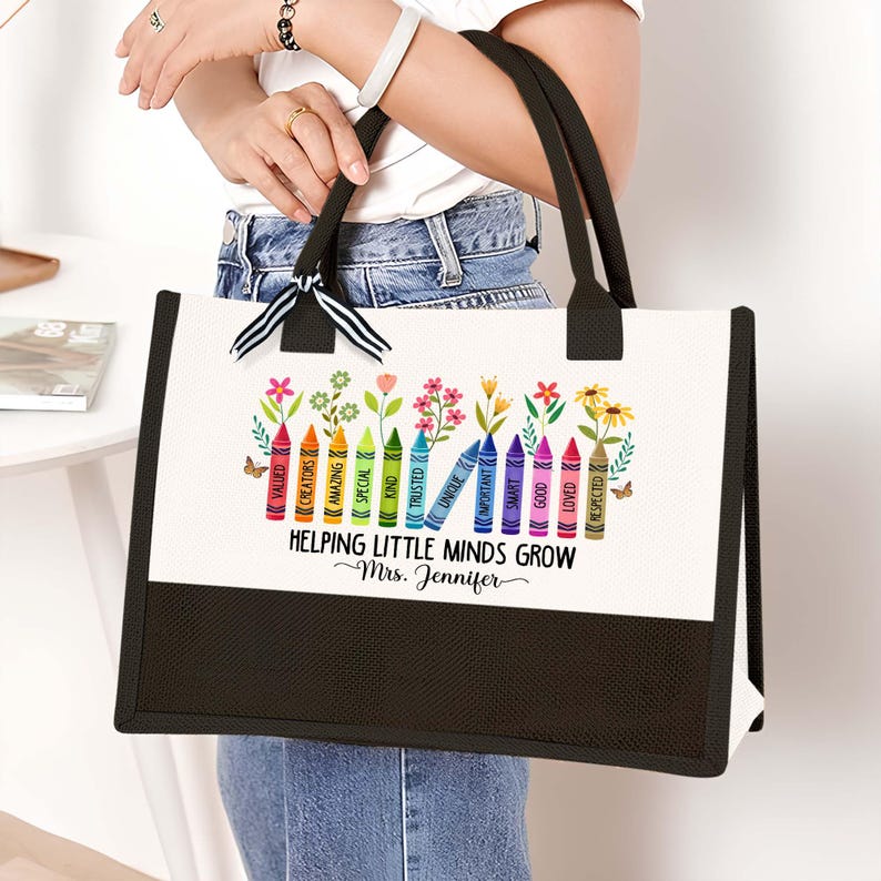 Puede incluir: Una bolsa de lona blanca con asas negras y fondo negro. La bolsa tiene un dise&ntilde;o colorido de l&aacute;pices de colores con palabras como "valued", "creators", "amazing", "special", "kind", "trusted", "unique", "invincible", "smart", "important", "good", "loved" y "respected". El texto "Helping Little Minds Grow" est&aacute; impreso debajo de los l&aacute;pices de colores, seguido de "~Mrs. Jennifer".