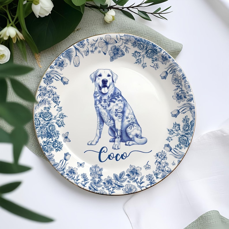 May include: A white ceramic plate with a blue floral border and a gold rim. The plate features a blue illustration of a Labrador Retriever dog with the name "Coco" written below. The plate is surrounded by greenery and white flowers.
