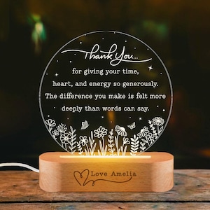 May include: A circular clear acrylic plaque with white engraved text reading Thank You for giving your time, heart, and energy so generously. The difference you make is felt more deeply than words can say, featuring floral and butterfly motifs, set in a light wood base with the engraved text Love Amelia.