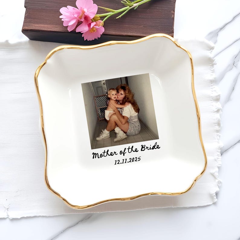 Op de afbeelding: Witte vierkante keramische schaal met een gouden rand en een golvende rand. De schaal heeft een foto van een moeder en kind, met de tekst "Mother of the Bride" en de datum 12.11.2025. Roze bloemen en een houten doos staan op de achtergrond.