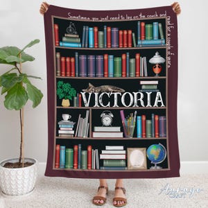 May include: A maroon fleece blanket with a bookshelf design featuring a cat sleeping on a shelf. The bookshelf is filled with books and other decorative items. The text "Sometimes you just need to lay on the couch and read for a couple of years" is at the top of the blanket. The name "VICTORIA" is written in white on a black shelf.