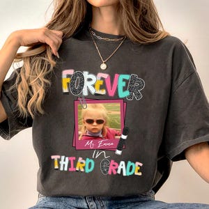 May include: A charcoal gray t-shirt featuring a graphic print with the text "FOREVER in THIRD GRADE". The design includes a photograph of a child wearing sunglasses inside a pink frame, accented by a paperclip, a pencil, and the name "Ms. Emma".
