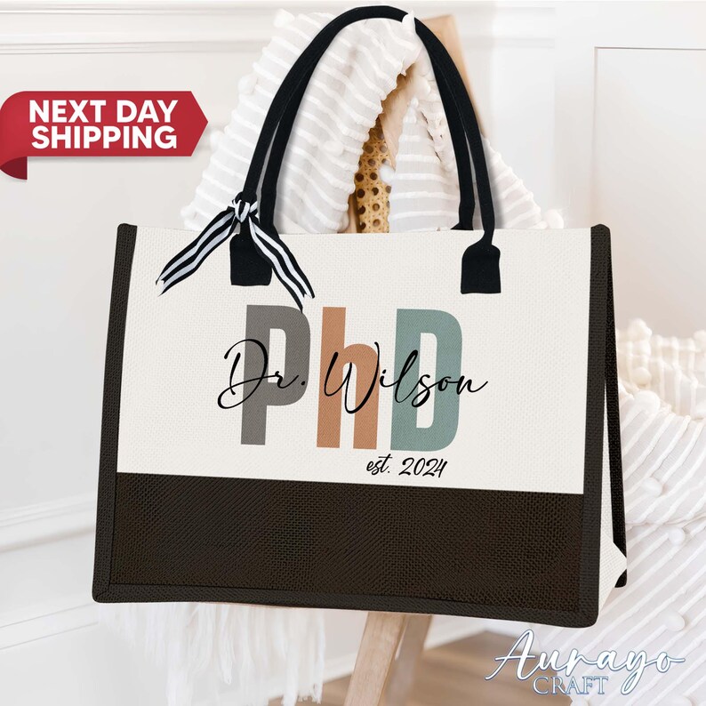 Pode incluir: Uma bolsa tote de lona branca com al&ccedil;as pretas e uma fita listrada preta e branca. A bolsa tem um fundo preto e o texto "Dr. Wilson PhD est. 2024" em letras coloridas.
