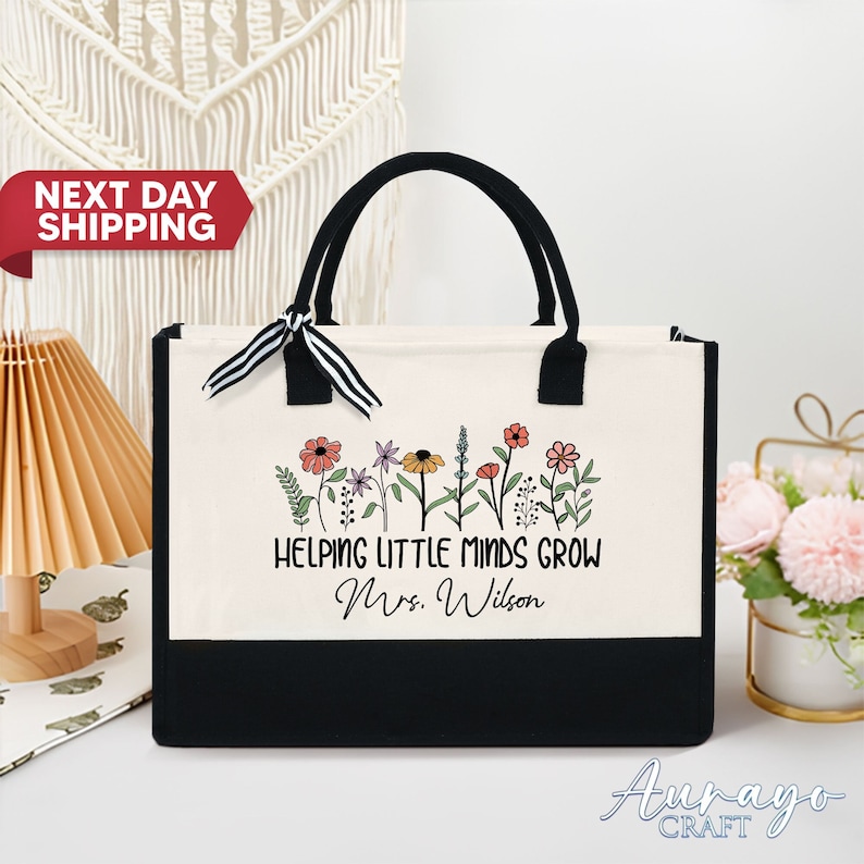 May include: A white canvas tote bag with black handles and a black bottom. The bag has a black and white striped ribbon tied around the handles. The bag has a floral design and the text "Helping Little Minds Grow Mrs. Wilson" printed on the front.