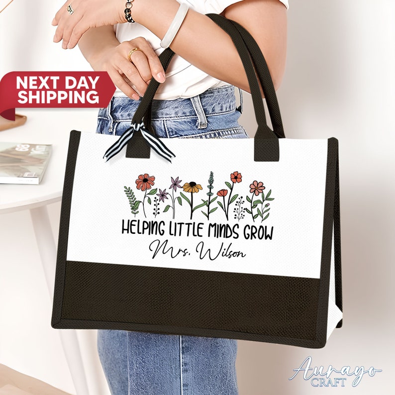 May include: White tote bag with black handles and a black bottom. The bag has a floral design with the text "Helping Little Minds Grow Mrs. Wilson".